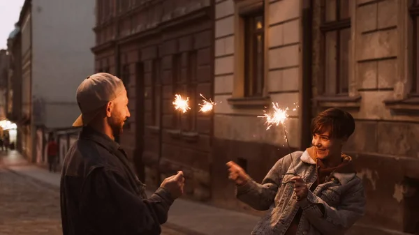 Young Stylish Couple Walking Through The City Street. In Their Hands The Burning Sparklers.