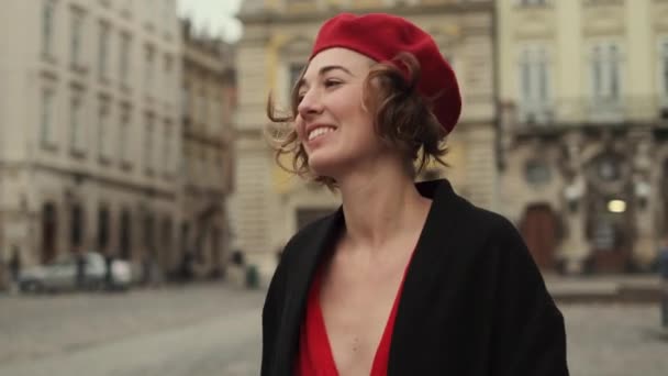 Mujer joven sonriente en boina roja y vestido rojo caminando en la plaza de la ciudad. — Vídeos de Stock