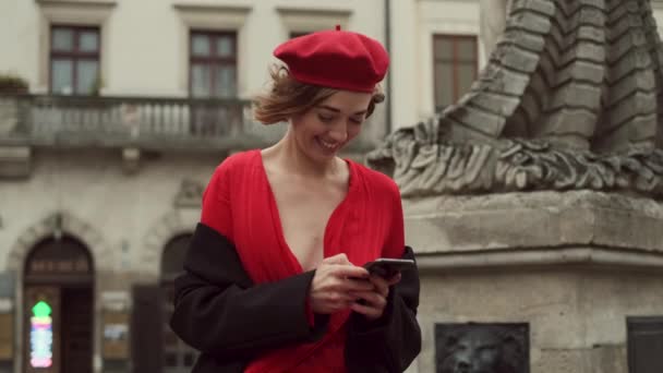 携帯電話を手に街の真ん中に立つ赤でスタイリッシュな若い女性. — ストック動画