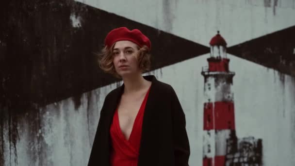 Młoda kobieta w płaszczu i czerwonej sukience stoi przy ścianie z obrazem latarni morskiej. Pretty Woman z krótkimi kręconymi włosami. — Wideo stockowe