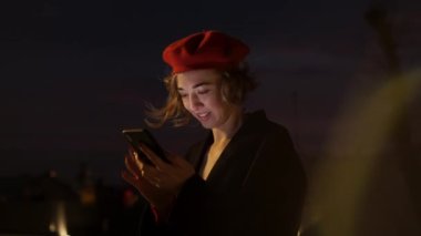 Alacakaranlık. Elinde cep telefonuyla caddenin ortasında dikilen Kırmızılı Şık Genç Kadın.