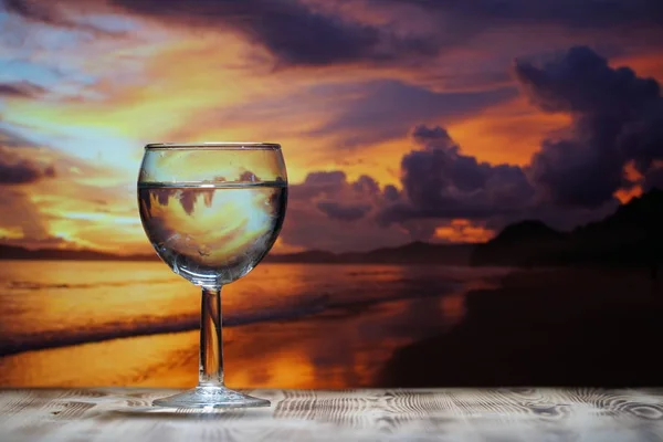 Ett glas dricksvatten är på ett träbord mot bakgrund av havet solnedgången. — Stockfoto