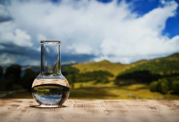Clean water in a glass laboratory flask on wooden table on mountain background. Ecological concept, the test of purity and quality of water.