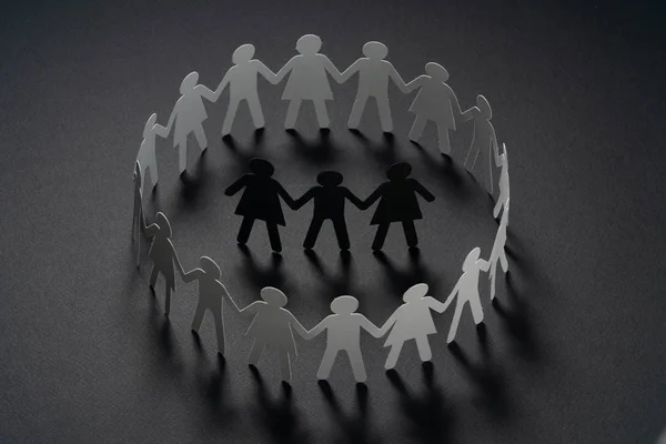 Three human paper figures surrounded by circle of paper people holding hands on dark surface. Bulling, segregation, conflict concept. — Stock Photo, Image