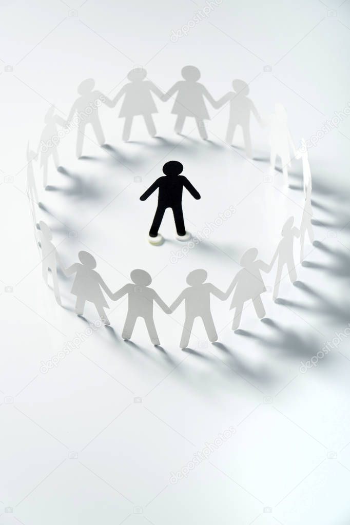 Paper human figure surrounded by circle of paper people holding hands on white surface. Bulling, segregation, conflict concept.