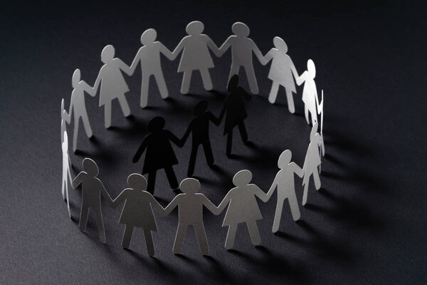 Three human paper figures surrounded by circle of paper people holding hands on dark surface. Bulling, segregation, conflict concept.