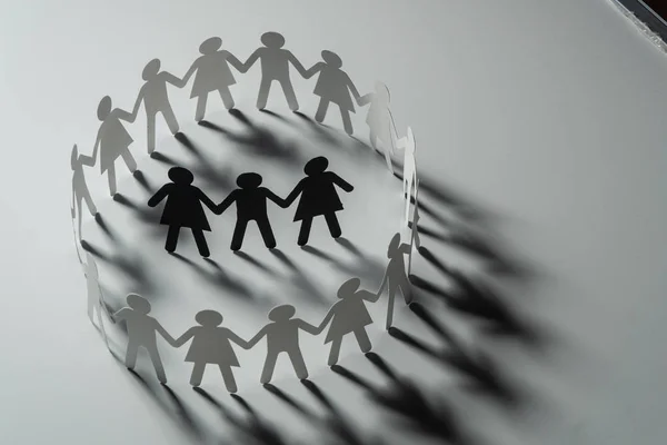 Three human paper figures surrounded by circle of paper people holding hands on white surface. Bulling, segregation, conflict concept. — Stock Photo, Image