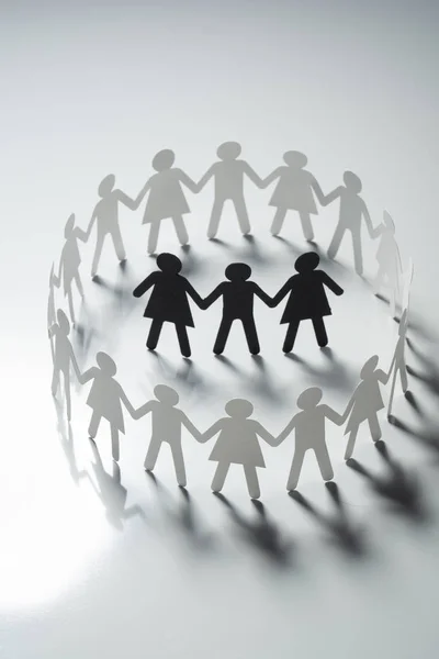 Three human paper figures surrounded by circle of paper people holding hands on white surface. Bulling, segregation, conflict concept. — Stock Photo, Image