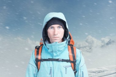 Kışın dağın tepesindeki genç dağcı erkek kameraya bakıyor. Ön manzara. Soğuk havada aktif bir yaşam tarzı. Kış dağı manzarası ve bulutlu gökyüzü.