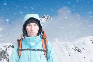 Kışın dağın tepesindeki genç dağcı erkek kameraya bakıyor. Ön manzara. Soğuk havada aktif bir yaşam tarzı. Kış dağı manzarası ve bulutlu gökyüzü.