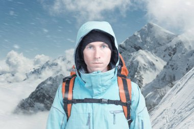 Kışın dağın tepesindeki genç dağcı erkek kameraya bakıyor. Ön manzara. Soğuk havada aktif bir yaşam tarzı. Kış dağı manzarası ve bulutlu gökyüzü.