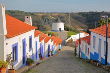 A row of colorful houses along a steep cobbled street inside Odeceixe (near Aljezur), leading to a whitewashed traditional windmill, Costa Vicentina, Algarve, Portugal clipart