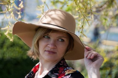 Genç sarışın, mavi gözlü, kahverengi şapkalı ve renkli elbiseli bir kadın parkta poz veriyor.