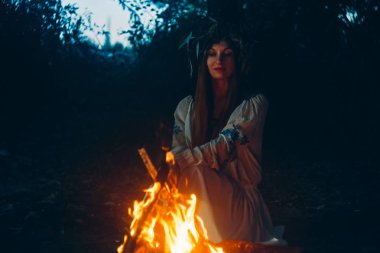 Kadın etnik kıyafetler içinde chaplet ile bir baş şenlik ateşi yakın oturuyor. Pagan tatil konsepti.