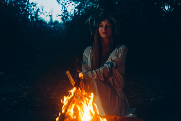 Woman in ethnic clothes with chaplet on a head sitting near bonfire. Concept of Pagan holiday.