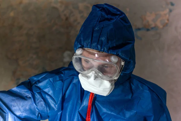 A man in blue protective suit, glasses and respirator look down