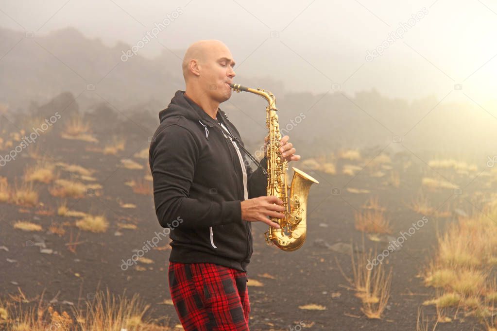 A bald man plays on a gold alto saxophone in the nature, against