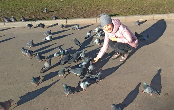 Молодая девушка улыбается и кормит стаю серых голубей на улице — стоковое фото