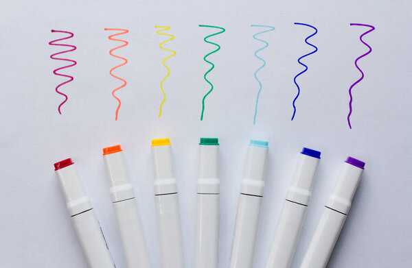 A set of bright multi-colored felt-tip pens or markers lie on white background sheet of paper, with samples of drawing. Creativity, drawing for children, design. Many Markers of all colors of rainbow.