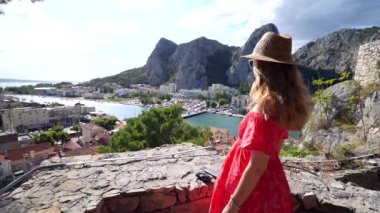 Uzun sarı saçlı, kırmızı elbiseli ve hasır şapkalı genç bir kız, kulenin tepesinde duruyor, gülümsüyor, Hırvatistan 'ın Omis kentine bakıyor ve eski kent, nehir ve köprünün manzarasını seyrediyor. Yavaş çekim.