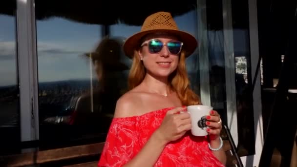 金髪の若い女の子は 赤いドレス 麦藁帽子とサングラスで 夏のカフェでスイングし 白い磁器のカップからコーヒーや紅茶を飲みます リラックス リラックス 気分が良い — ストック動画