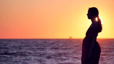 Elbiseli kız gölgesi denizin arka planında duruyor, güzel bir gün batımı, gün doğumu. Yavaş çekim videosu. Rüzgarda çırpınan elbiseler, deniz ve günbatımının ışınlarında parlayan dalgalar ve güneşte yanan ışıklar..