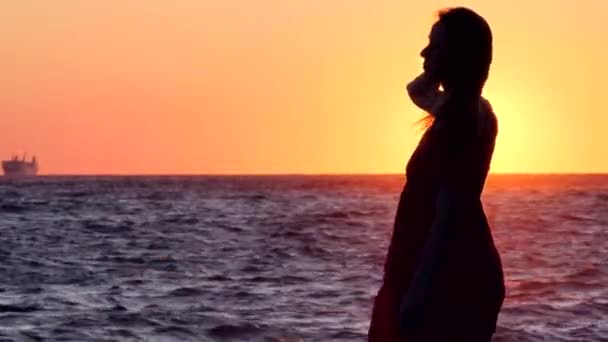 穿着衣服的女孩轮廓站在大海的背景上 美丽的日落 衣服在风中飘扬 海浪在夕阳中闪耀 在阳光中燃烧 女孩摸头发 玩头发 — 图库视频影像