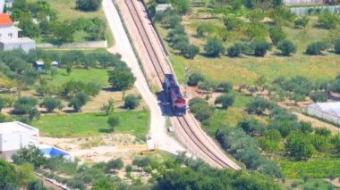Hırvatistan, Split, 19 Ağustos 2019. İki vagonluk bir tren, mavi-kırmızı-beyaz renkte, Split kenti yakınlarındaki demiryollarıyla, konut binaları ve bahçeler arasında, yazın seyahat eder..