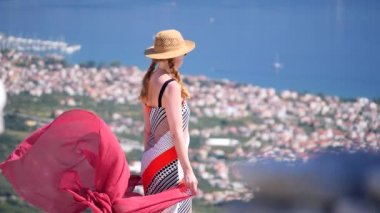 Açık kahverengi örgülü ve hasır şapkalı genç bir kız, Hırvatistan 'ın Split kentindeki dağın tepesinde rüzgarda dalgalanan kırmızı bir eşarp tutuyor. Kız hayatı, rahatlamayı ve rüzgarı seviyor. Yavaş çekim.