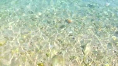 Taşlarla kaplı güzel bir deniz yatağı. Turkuaz mavi deniz suyu güneşte sığ dalgalarda parıldar ve güneş parıltısı yaratır. Spa için deniz ekran koruyucusu, rahatlama. Tatil videosu, yaz tatili. Deniz arkaplanı.