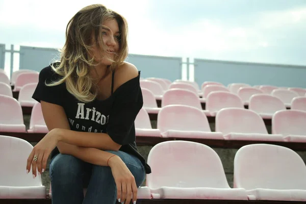 Urbana porträtt av en flörtig vacker kvinna i svart t-shirt och jeans på arenan, sitter på en bänk — Stockfoto