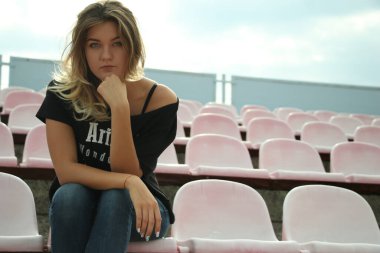 Şehir portresi, siyah tişörtlü ve kot pantolonlu güzel bir kadının stadyumda bankta oturması.