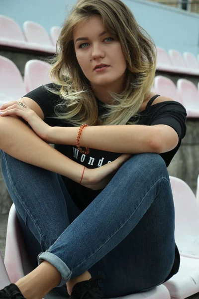 Stedelijk portret van een flirterige mooie vrouw in zwart t-shirt en jeans in het stadion, zittend op een bankje — Stockfoto