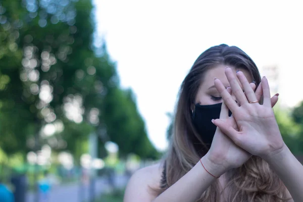 Mujer rubia bastante joven con máscara facial negra médica. Llevando una camiseta y pantalones cortos. en un parque. realidad moderna. concepto covid-19. espacio de copia . — Foto de Stock