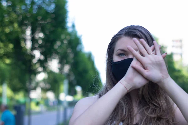 Pretty young blonde woman in medical black face mask. Wearing a t-shirt and jeans shorts. in a park. modern reality. covid-19 concept. copy space. — Stock Photo, Image