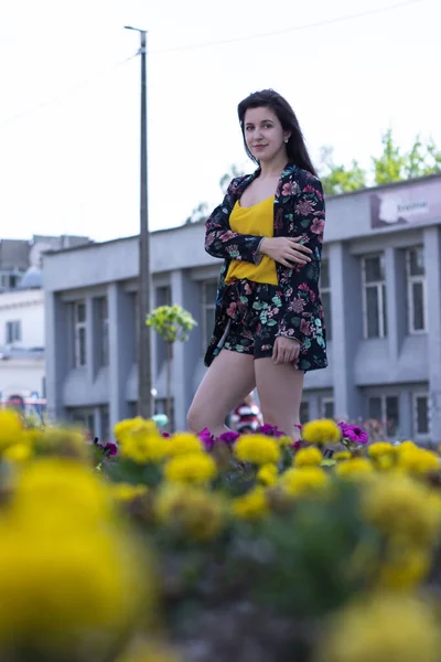 Vonzó fiatal barna nő sárga pólóban és virágos rövidnadrág jelmezben. közel egy bokor virághoz. másolás helye. — Stock Fotó