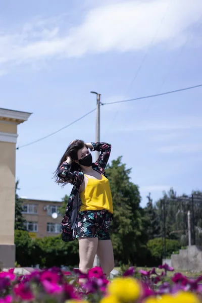 Ελκυστική νεαρή μελαχρινή γυναίκα με κίτρινο μπλουζάκι και λουλουδάτη φορεσιά σορτς. Με ιατρική μαύρη μάσκα προσώπου. σύγχρονη πραγματικότητα. κοντά σε θάμνους με λουλούδια. Έννοια covid-19. αντίγραφο χώρου. — Φωτογραφία Αρχείου