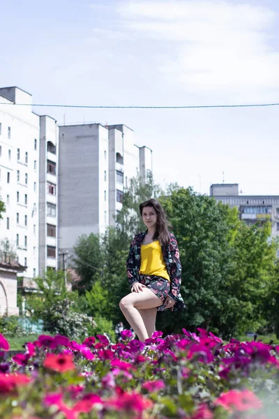 Ελκυστική νεαρή μελαχρινή γυναίκα με κίτρινο μπλουζάκι και λουλουδάτη φορεσιά σορτς. κοντά σε ένα θάμνο με λουλούδια. αντίγραφο χώρου. — Φωτογραφία Αρχείου