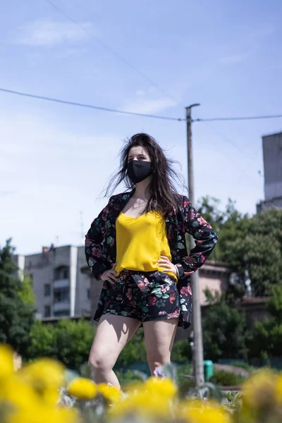 Mulher morena jovem atraente em camiseta amarela e traje de shorts floridos. Em máscara facial preta médica. realidade moderna. perto de arbustos com flores. conceito covid-19. espaço de cópia . — Fotografia de Stock