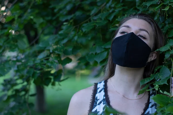 Mujer rubia bastante joven con máscara facial negra médica. en un arbusto, bosque. concepto covid-19. realidad moderna. espacio de copia . — Foto de Stock