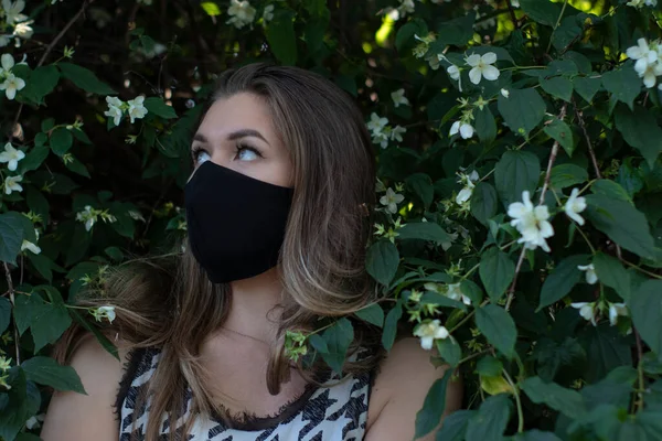 Pretty young blonde woman in medical black face mask. in a bush with little white flowers. modern reality. covid-19 concept. copy space. — Stock Photo, Image