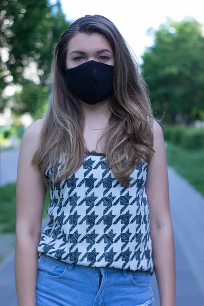 Pretty young blonde woman in medical black face mask. Wearing a t-shirt and jeans shorts. in a park. modern reality. covid-19 concept. copy space. — Stock Photo, Image