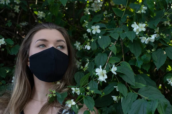 Bella giovane donna bionda in maschera medica nera. in un cespuglio con piccoli fiori bianchi. realtà moderna. covid-19 concetto. copia spazio . — Foto Stock