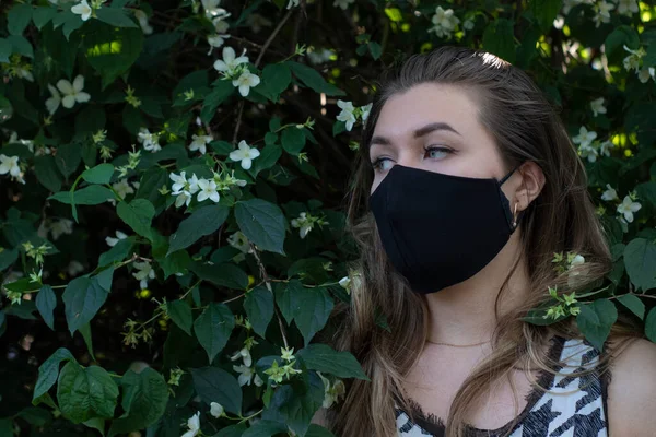 Mujer rubia bastante joven con máscara facial negra médica. en un arbusto con pequeñas flores blancas. realidad moderna. concepto covid-19. espacio de copia . — Foto de Stock