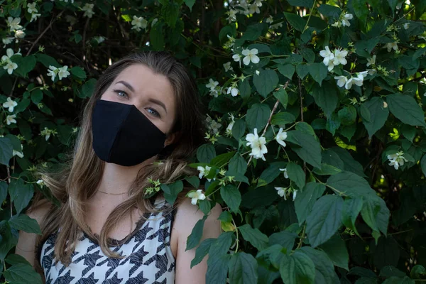 Hübsche junge blonde Frau mit medizinischer schwarzer Gesichtsmaske. in einem Busch mit kleinen weißen Blüten. Moderne Realität. covid-19-Konzept. Kopierraum. — Stockfoto