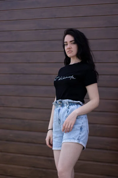 Bela mulher morena atraente em t-shirt preta e shorts jeans em um fundo de parede marrom — Fotografia de Stock