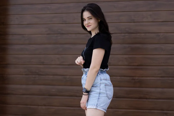 Schöne attraktive brünette Frau in schwarzem T-Shirt und Jeans-Shorts auf einem Hintergrund aus brauner Wand — Stockfoto