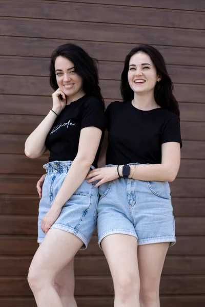 Dos mujeres morenas jóvenes con una camiseta negra y pantalones cortos de mezclilla abrazándose. Hermanas, novias.familia y concepto de amistad —  Fotos de Stock