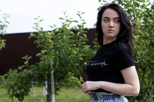迷人的年轻黑发女人穿着黑色T恤和牛仔短裤。在乡下，在花园里. — 图库照片