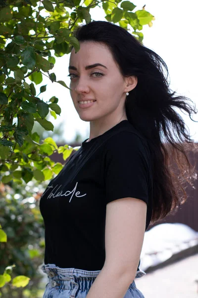 검은 티셔츠에 데님 반바지입은 젊은 갈색 머리 여자를 소개 합니다. 시골에서, 정원에서. — 스톡 사진